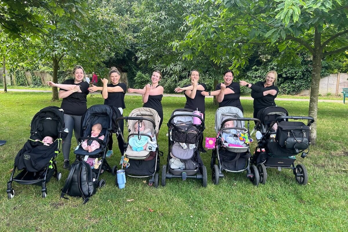 A group of mums from an NCT group, accompanied by thier babies, doing a shoulder stretch in a personal training session